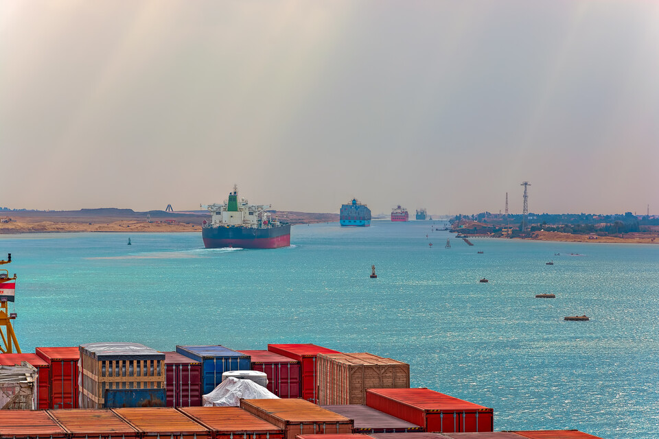 Im Vordergrund sind mehrere Container zu sehen. Im Hintergrund fahren durch einen Handelskanal mehrere Containerschiffe.
