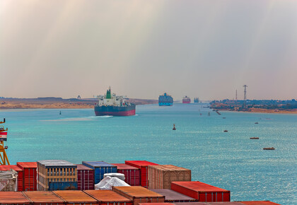 Im Vordergrund sind mehrere Container zu sehen. Im Hintergrund fahren durch einen Handelskanal mehrere Containerschiffe.