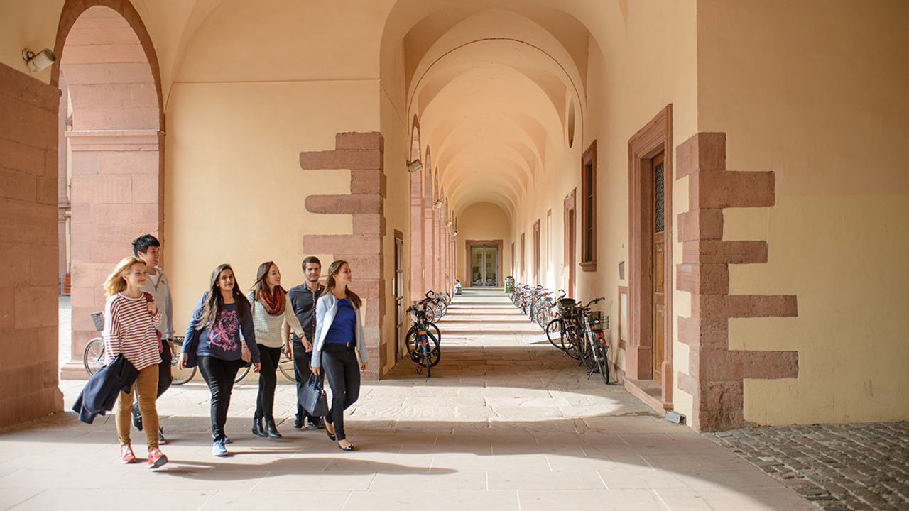 Vier Studentinnen und zwei Studenten spazieren über den Campus der Universität Mannheim. Die Studierenden sind frühlingshaft gekleidet. In einem Gang im Hintergrund stehen viele Fahrräder an den sandfarbenen Wänden des Schlosses.