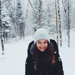 Eine junge Frau trägt eine schwarze Winterjacke und eine graue Mütze. Im Hintergrund ist ein schneebedeckter Wald zu sehen.