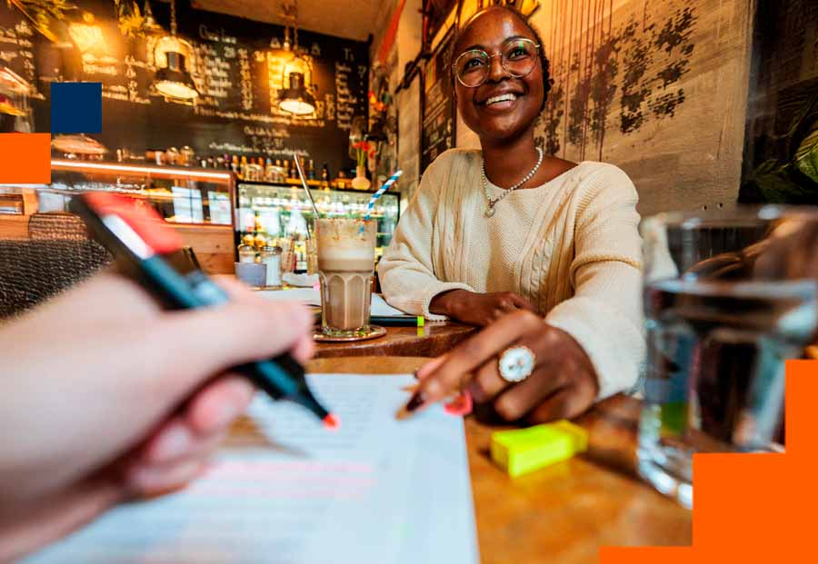 Eine Studentin sitzt in einem Café, vor ihr steht ein Café und ihr gegenüber sitzt eine Person, von der nur die Hand mit einem Textmarker darin zu sehen ist.