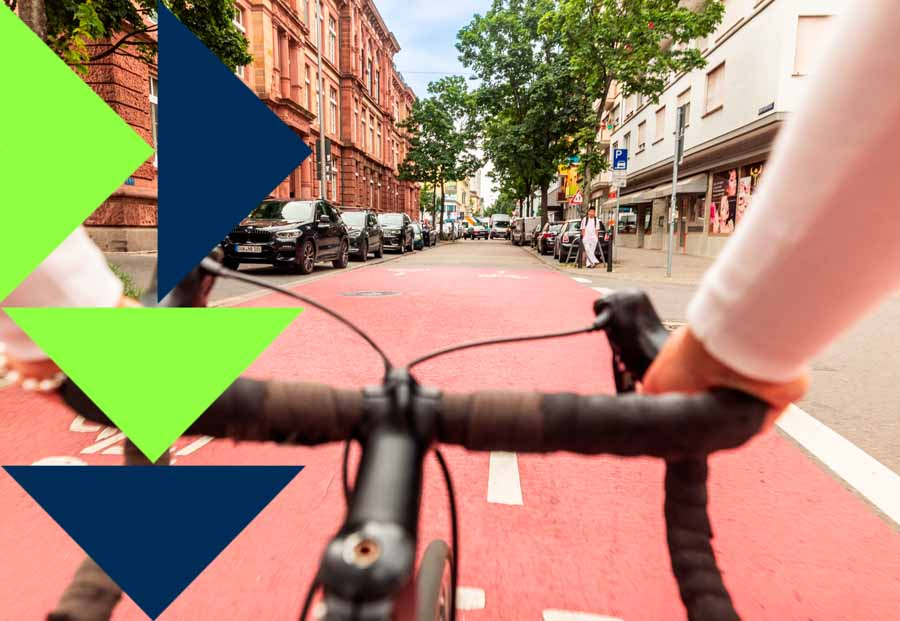 Der Lenker eines Fahrrads wird im Vordergrund von zwei Händen gegriffen, das Fahrrad fährt auf einer Fahrradstraße, im Hintergrund Gebäude und Bäume.