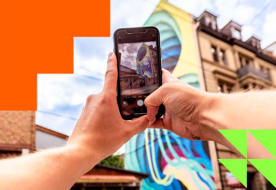 Zwei Hände halten ein Handy, das wiederum ein Foto von einem bunten Mural an einer Hauswand macht.