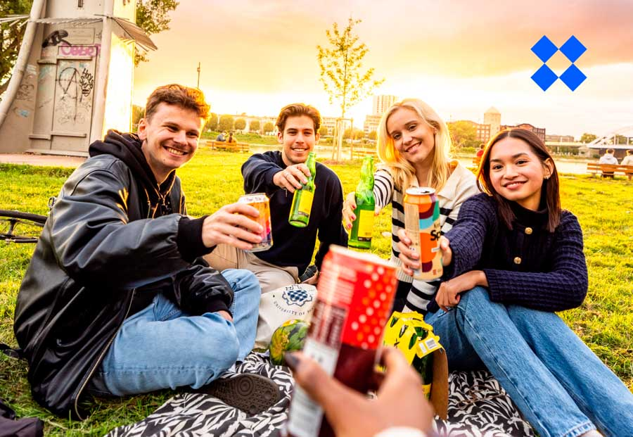 Zwei Studentinnen und zwei Studenten sitzen auf einer Picknickdecke auf einer Wiese. Sie stoßen mit der Person hinter der Kamera an, deren Hand mit einer Getränkedose noch zu sehen ist.