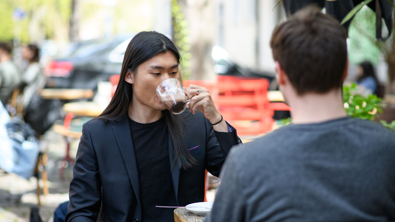 Ein Student mit glatten langen schwarzen Haaren und Jacket trinkt Kaffee aus einem Glas. Er sitzt draußen in einem Café.