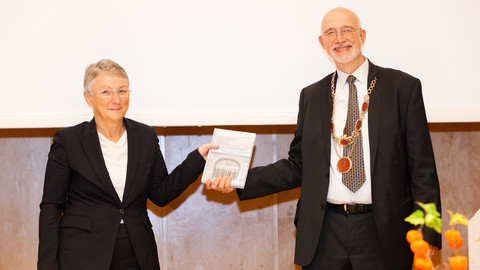 Zwei lächelnde Personen in schicker Kleidung stehen nebeneinander und halten die Festschrift zum 75-jährigen Jubiläum der Neubegründung in den Händen. Die Personen heißen Barbara Windscheid und Thomas Puhl.