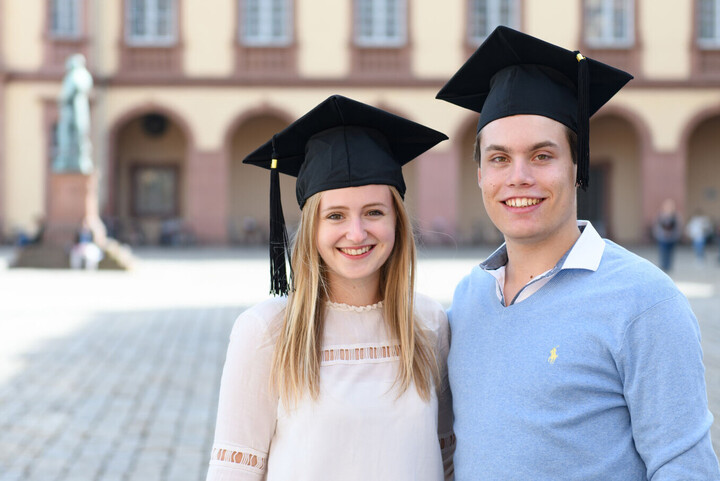 Eine Bachelorabsolventin und ein Bachelorabsolvent stehen vor dem Mannheimer Schloss.