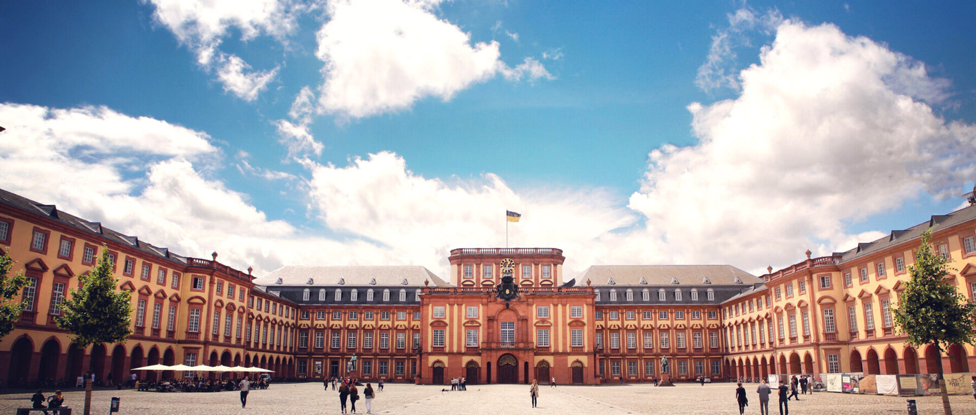 Das Barock-Schloss und der Ehrenhof der Universität Mannheim unter strahlend blauem Himmel. Das Schloss ist von unzähligen Fenstern, rotem Sandstein und einer gelben Fassade geprägt.