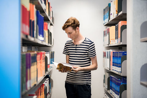 Ein Student hält ein aufgeschlagenes Buch in seinen Händen und blättert darin. Neben ihm stehen gut gefüllte Bücherregale. Der Student trägt kurzes blondes Haar und ein schwarz-weiß gestreiftes T-Shirt.