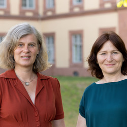 Ursula Schlichter und Silvia Luber stehen vor dem Mannheimer Schloss. Ursula Schlichter hat graue schulterlange Haare und trägt eine rostrote Bluse. Silvia Luber hat rötlich-braune Haare und trägt ein blaues Oberteil. 