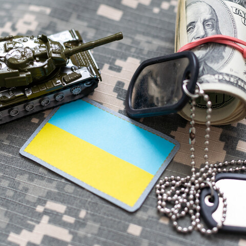 Auf einem Tisch liegen ein Spielzeugpanzer, eine kleine ukrainische Flagge, Dollarscheine und zwei militärische Erkennungsmarken ohne Schrift.