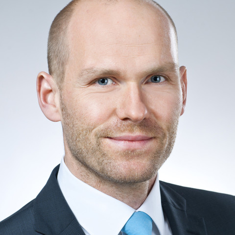 Eine Person trägt ein weißes Hemd, eine blaue Krawatte sowie einen dunkelblauen Blazer und steht vor einem weißen Hintergrund. Die Person heißt Florian Kutzner.