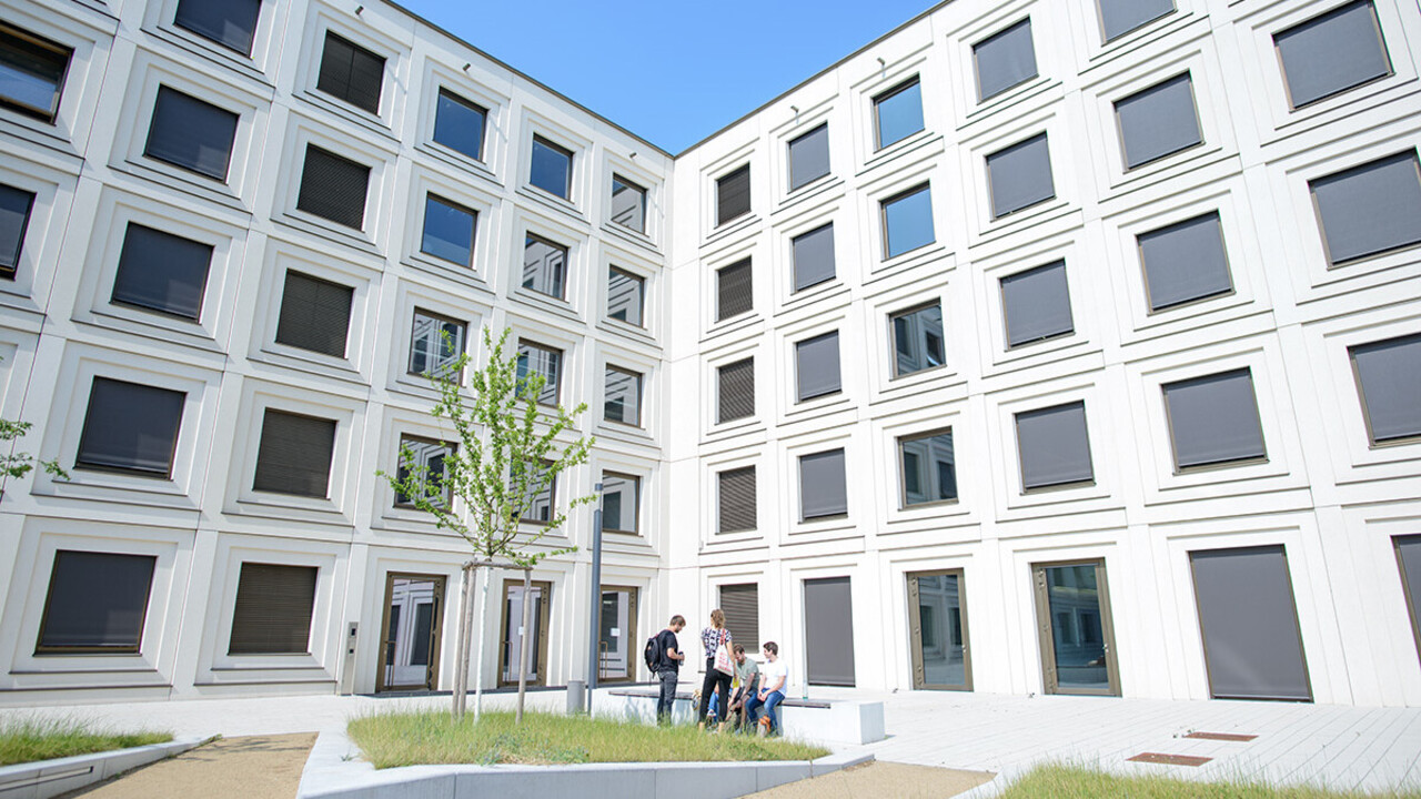 Innenhof des mit zahlreichen Fenstern gebauten Universitätsgebäudes B6 mit kleinen Grünflächen und im Hintergrund sitzen Studierende auf einer Bank.