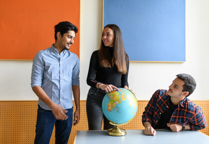 Neben zwei stehenden Personen sitzt eine Person an einem Tisch. Die Person in der Mitte zeigt auf einen Globus, der auf dem Tisch steht.
