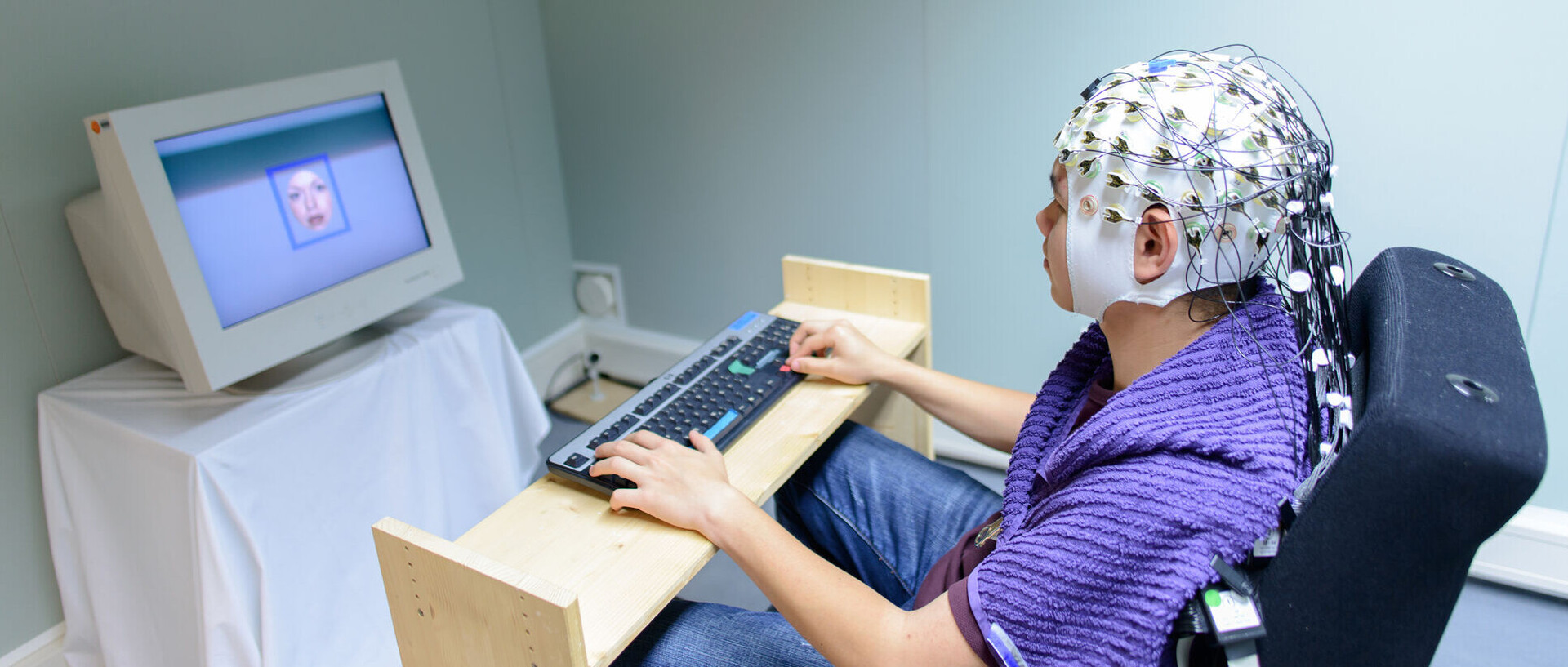 Eine Person trägt eine Haube mit Elektroden auf dem Kopf. Sie sitzt an einer Tastatur vor einem Monitor.