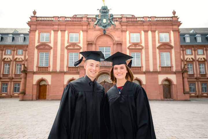 Zwei Studierende mit schwarzer Robe und Doktorhut stehen vor dem Schloss der Universität Mannheim