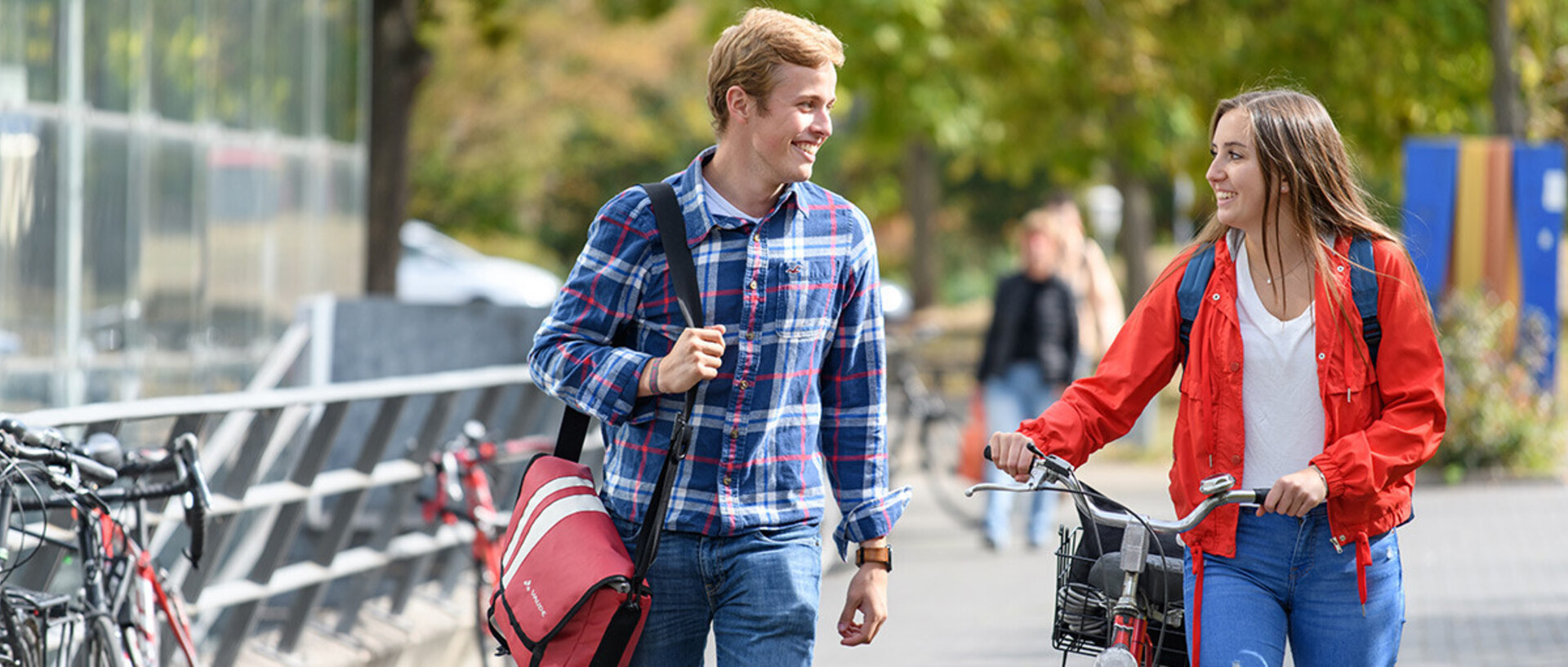 Eine Studentin und ein Student laufen lachend nebeneinander auf einem gepflasterten Weg. Die Studentin schiebt ihr Rad neben sich her.