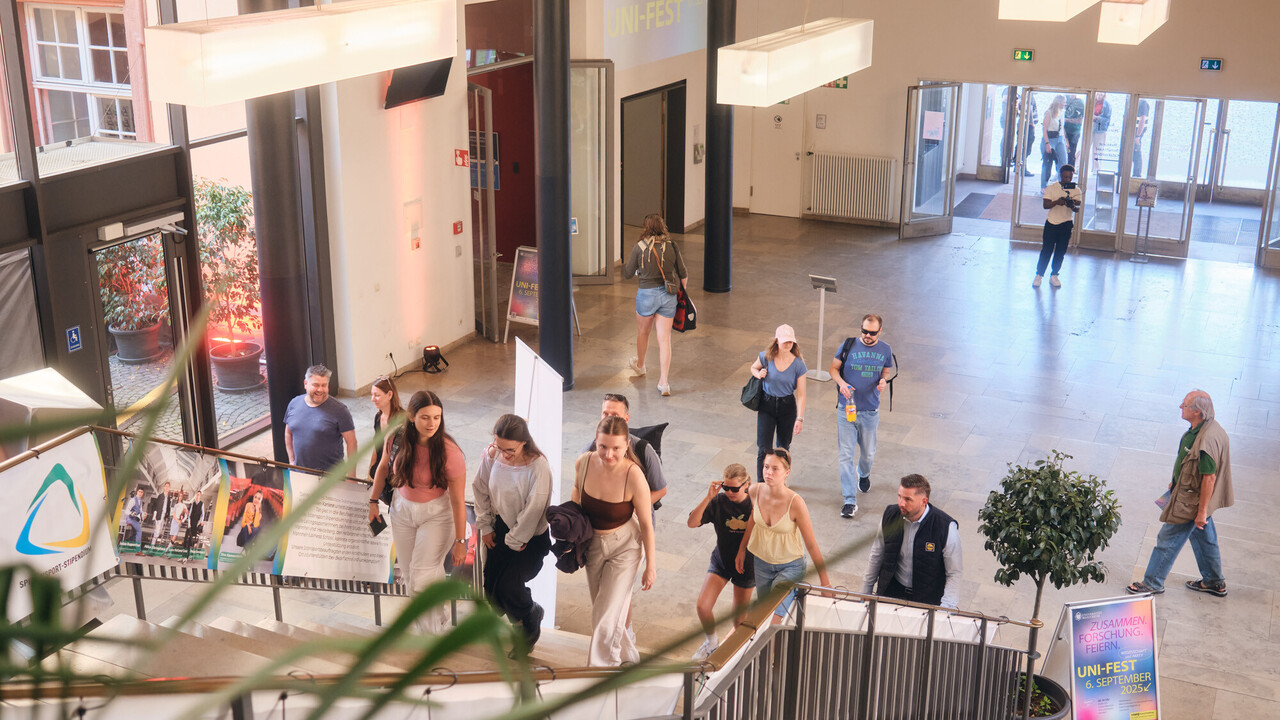 Personen steigen die Treppe im Foyer des Ostflügels der Uni Mannheim hoch, im Hintergrund sind Plakate und eine Projektion der Veranstaltung "Uni-Fest".