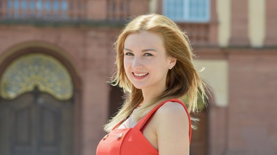 Eine lächelnde Person trägt ein orange-farbenes Top und steht vor dem Schloss der Universität Mannheim. Die Person heißt Katrina Cebatko.