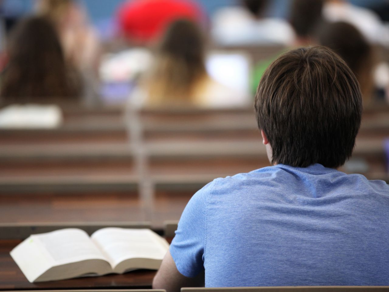 Rückenansicht eines Studierenden mit blauem T-Shirt und braunen Haaren. Er sitzt in einem Hörsaal, vor ihm ein aufgeklapptes Buch.