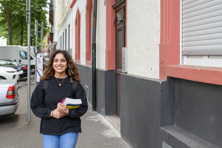 Eine Studierende lächelt und läuft mit einem Buch in der Hand durch eine Straße in Mannheim.
