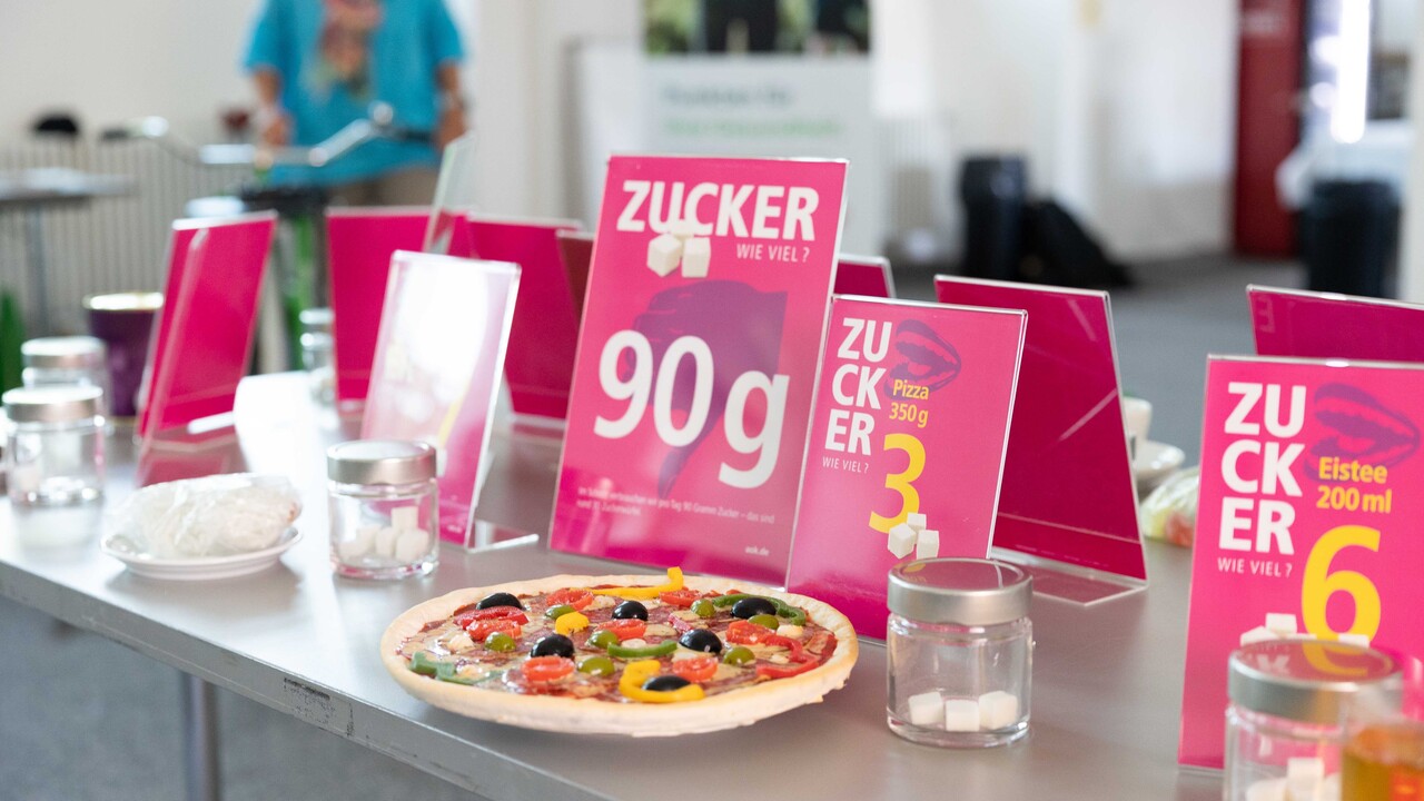 Verschiedenes Essen, darunter eine Pizza, ist auf Tellern an einem Tisch ausgestellt. Dahinter stehen Schilder mit Zuckerangaben.