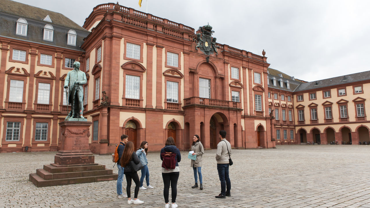 Vier Studierende hören einer Studentin beim Erzählen zu. Sie stehen auf dem Ehrenhof der Universität Mannheim. Im Hintergrund ist das Mannheimer Schloss zu sehen.