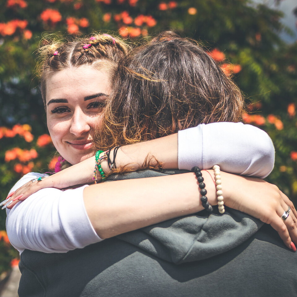 Zwei Personen umarmen sich. Eine Person steht mit dem Rücken zur Kamera, die andere Person lächelt.
