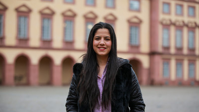 Eine lächelnde Person trägt eine lila-farbene Bluse sowie eine schwarze Jacke und steht vor dem Schloss. Die Person heißt Hala Fayad. 