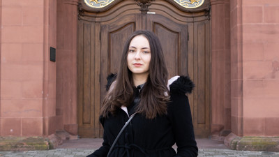 Eine Person trägt eine schwarze Jacke mit schwarzem Fell und steht vor einer braunen Schlosstüre. Die Person heißt Dijana Babic.