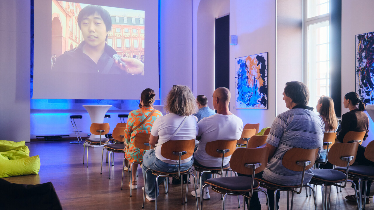 Zuschauer sitzen in Stuhlreihen vor einer Leinwand und schauen ein Video, in dem ein Mann am Schloss interviewt wird.
