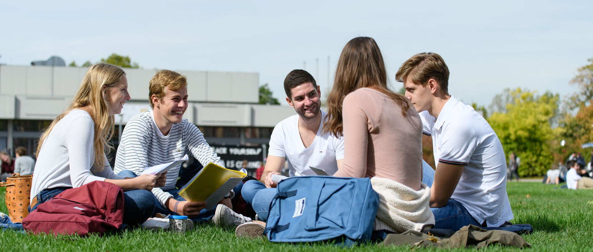 Eine Gruppe von fünf Studierenden sitzt gut gelaunt in einem Kreis auf der Mensawiese. In ihren Händen haben sie Blöcke oder Laptops, neben ihnen stehen Rucksäcke.