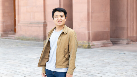 Eine lächelnde Person trägt einen grauen Pullover, eine blaue Jeans sowie eine braune Jacke und steht vor dem Schloss der Uni Mannheim. Die Person heißt Noel Chia.