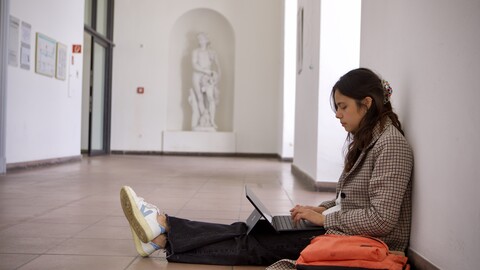 Eine studierende Person sitzt auf dem Boden des Schlosses an eine Wand gelehnt und schaut auf ihren Laptop.