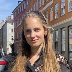 Eine langhaarige, blonde Studentin steht auf einer Straße und lächelt in die Kamera.