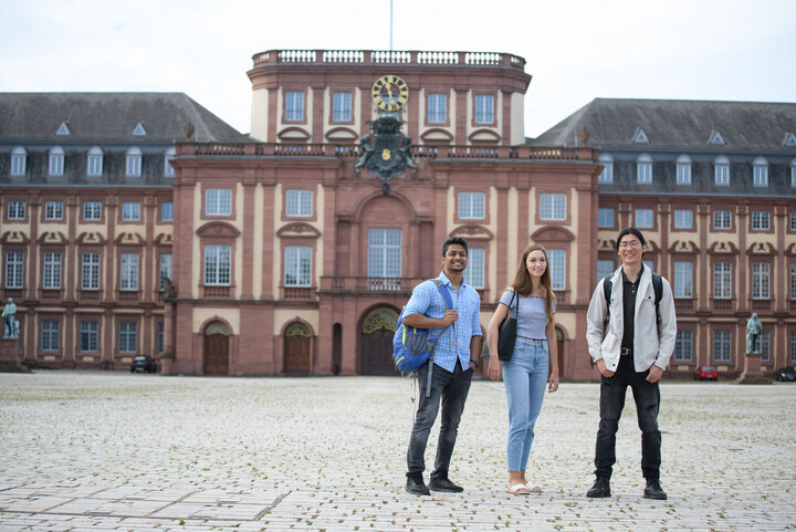 Drei Studierende auf dem Ehrenhof gucken in Richtung Kamera. Im Hintergrund der Mittelbau des Schlosses.