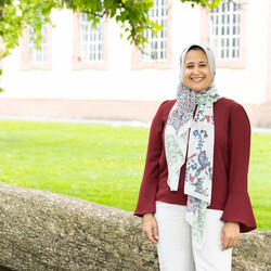 Alaa Elfawal stands smiling in front of a tree. She is wearing a colorful headscarf, a red blouse, and white trousers. The Schloss is in the background.