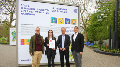 Vier Personen stehen vor einem Plakat mit den 17 Nachhaltigkeitszielen auf der Buga. Eine Studentin hält ein Zertifikat in den Händen.
