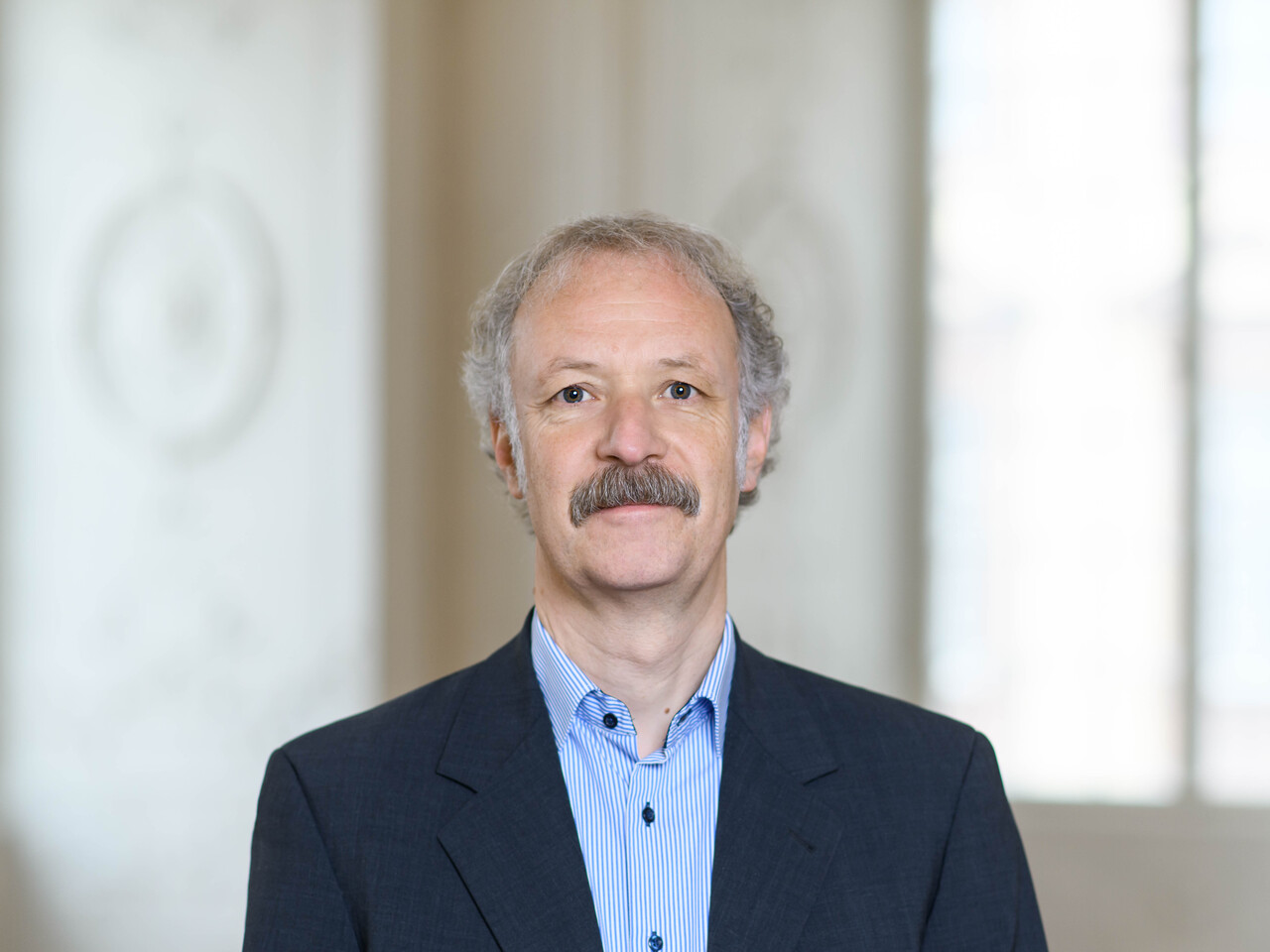 Prof. Dr. Moritz Fleischmann hat graue Haare und einen Schnauzer. Er trägt ein dunkles Jacket und ein gestreiftes Hemd.