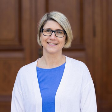 Eine lächelnde Person trägt ein blaues Top sowie einen weißen Blazer und steht vor einer braunen Holztüre. Die Person heißt Christina Hartmann.