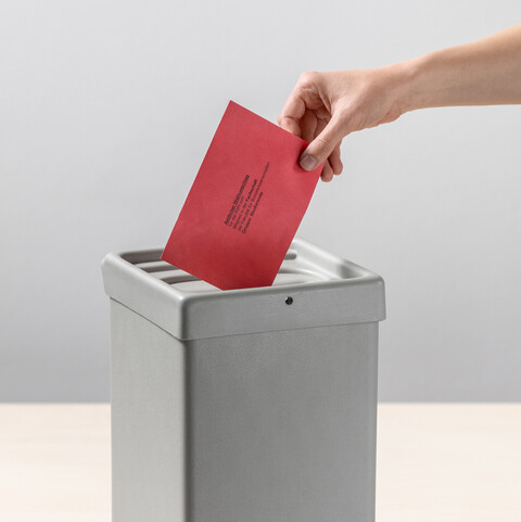 Eine Person steckt einen roten Umschlag in eine Wahlurne.