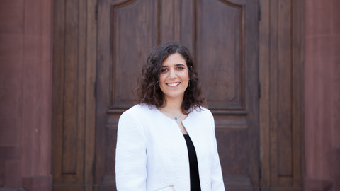 Eine lächelnde Person trägt ein schwarzes Top sowie einen weißen Blazer und steht vor einer Holztüre. Die Person heißt Amina Elbarbary.