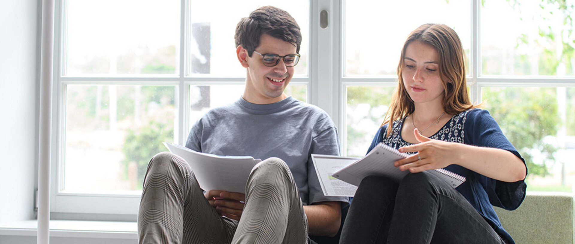Ein Student und eine Studentin sitzen in seinem Seminarraum am Fenster und betrachten gemeinsam Notizen.