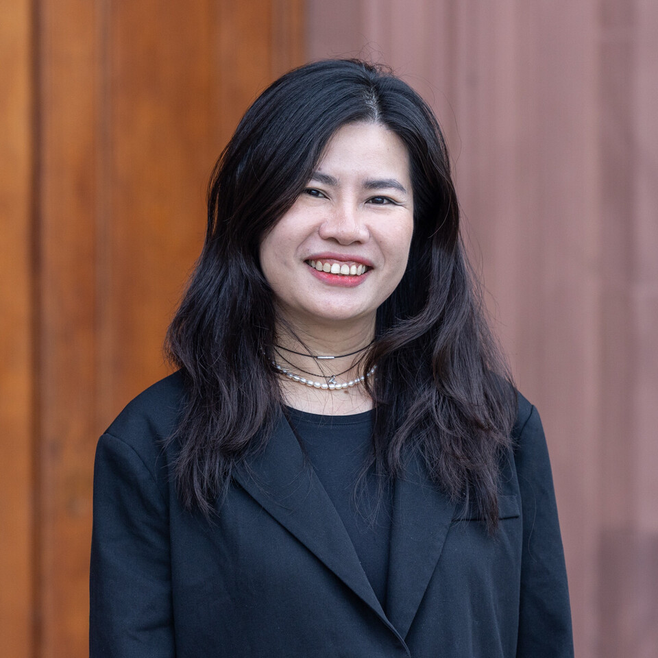 Porträt von Thuy Nguyen. Sie steht vor einem braunen Hintergrund, trägt ein schwarzes Jackett und lächelt in die Kamera.