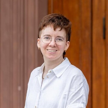 Anna Adendorf hat kurze braun-rote Haare und trägt eine Brille. Anna Adendorf hat helle Haut und leichte Sommersprossen.