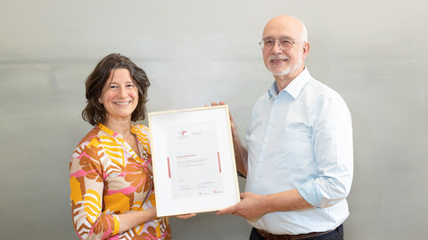 Kanzlerin Dr. Katrin Schoppa-Bauer und Rektor Professor Dr. Thomas Puhl halten ein Zertifikat in den Händen und lächeln in die Kamera.