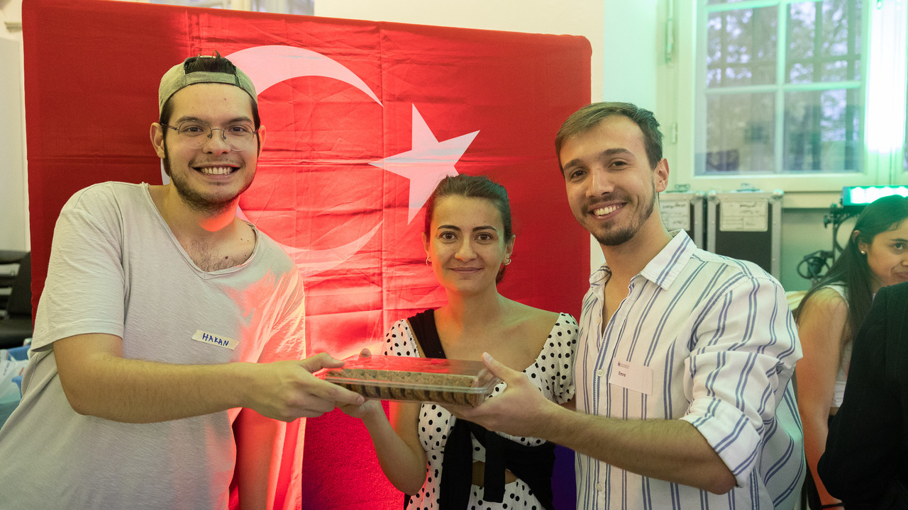 Drei Menschen stehen lächelnd vor einer türkischen Flagge und halten eine türkische Spezialität hoch.