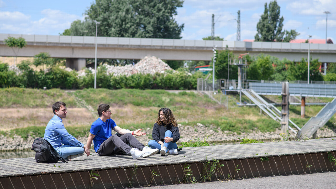 Drei Studierende sitzen in der Sonne auf einem Holzsteg. Im Hintergrund eine Brücke.