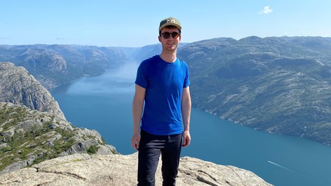 Eine Person in Wanderkleidung steht auf einem Felsvorsprung. Im Hintergrund ist ein See und Gebirge. Die Person heißt Timo Eberhard.