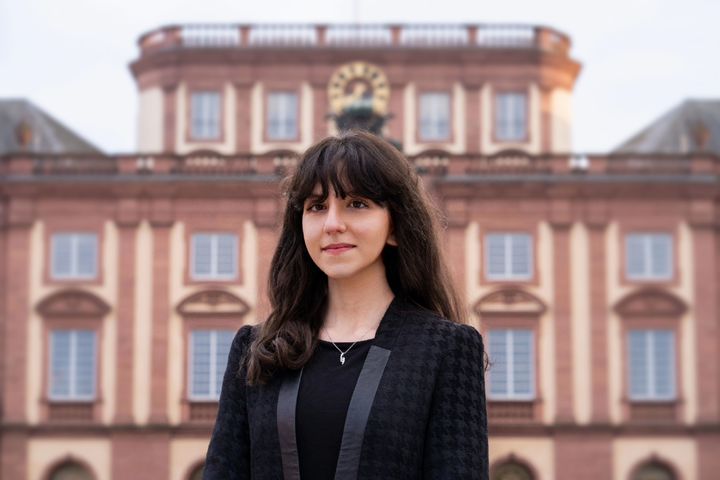 Eine Person trägt ein schwarzes Top sowie einen schwarz-grauen Blazerund steht vor dem Schloss Mannheim. Die Person heißt Nora Younis.